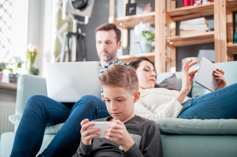 Familie entspannt im Wohnzimmer, jeder nutzt Smartphone, Tablet oder Laptop für digitale Medien.